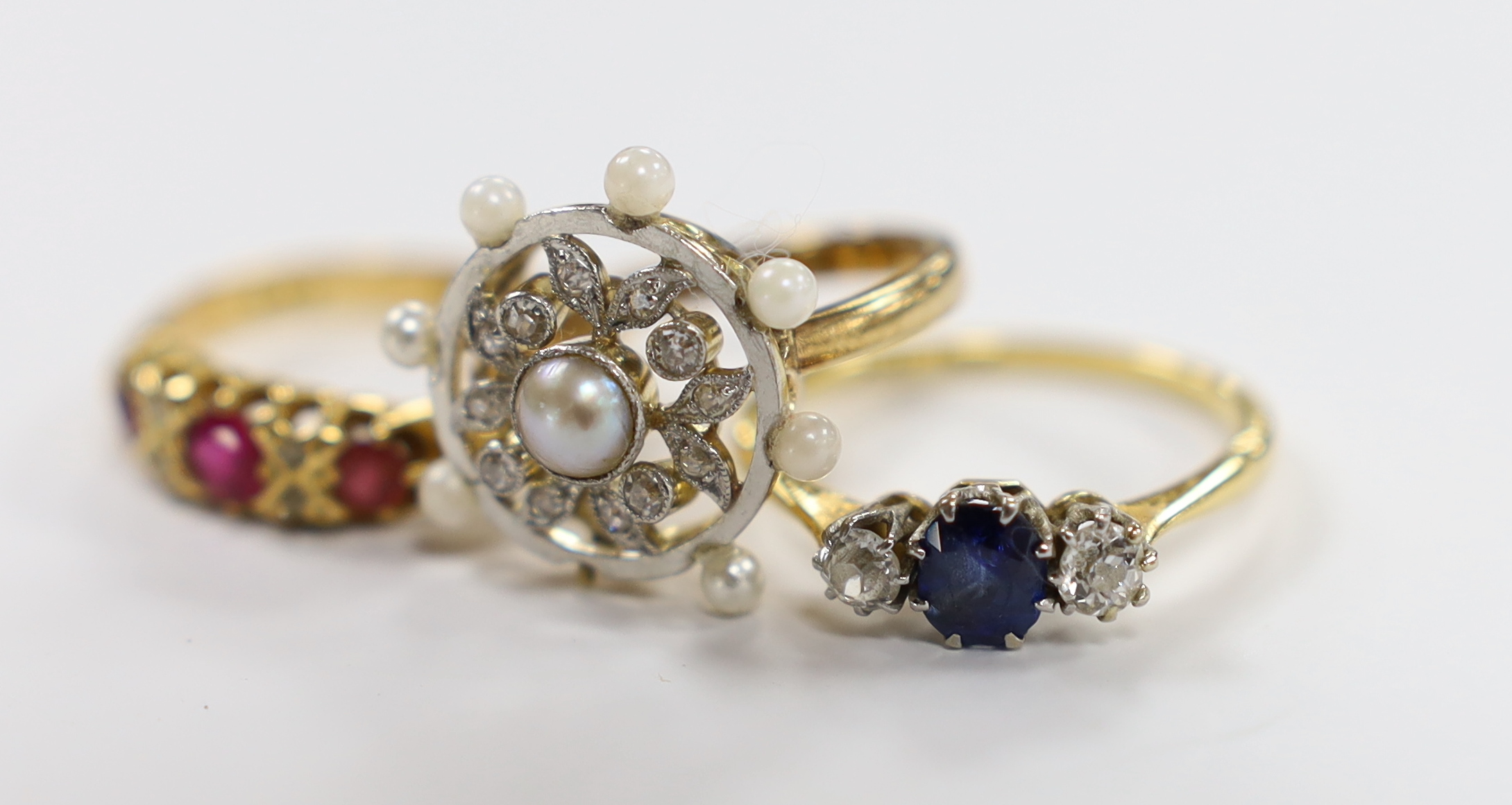 An 18ct, sapphire and diamond set three stone ring, a George V 18ct gold and three red stone set ring with diamond chip spacers and a yellow metal seed pearl and diamond chip set cluster ring (pearl missing), gross weigh
